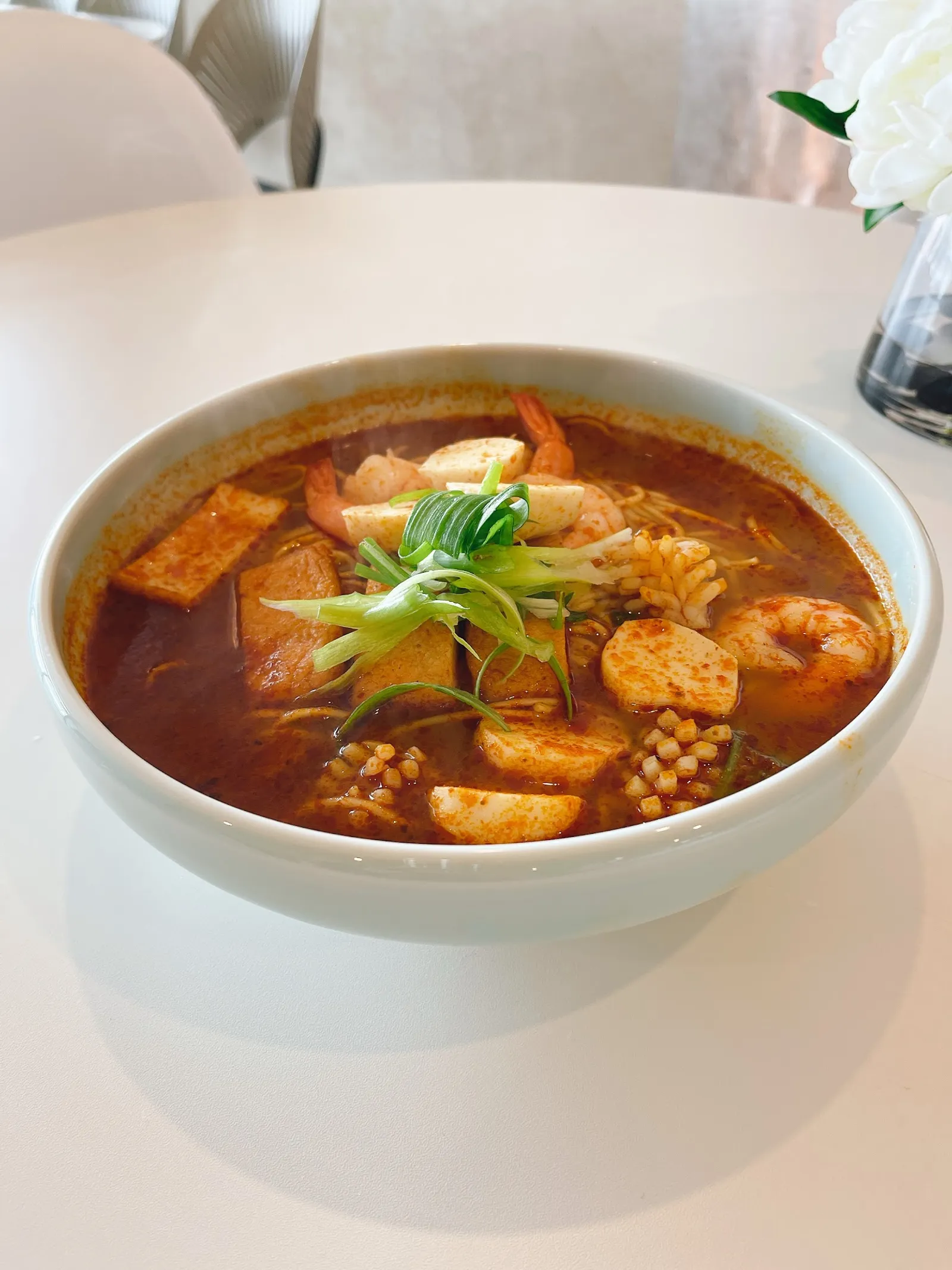 White bowl of tomyum curry noodle soup with prawns, tofu and spring onions on white table