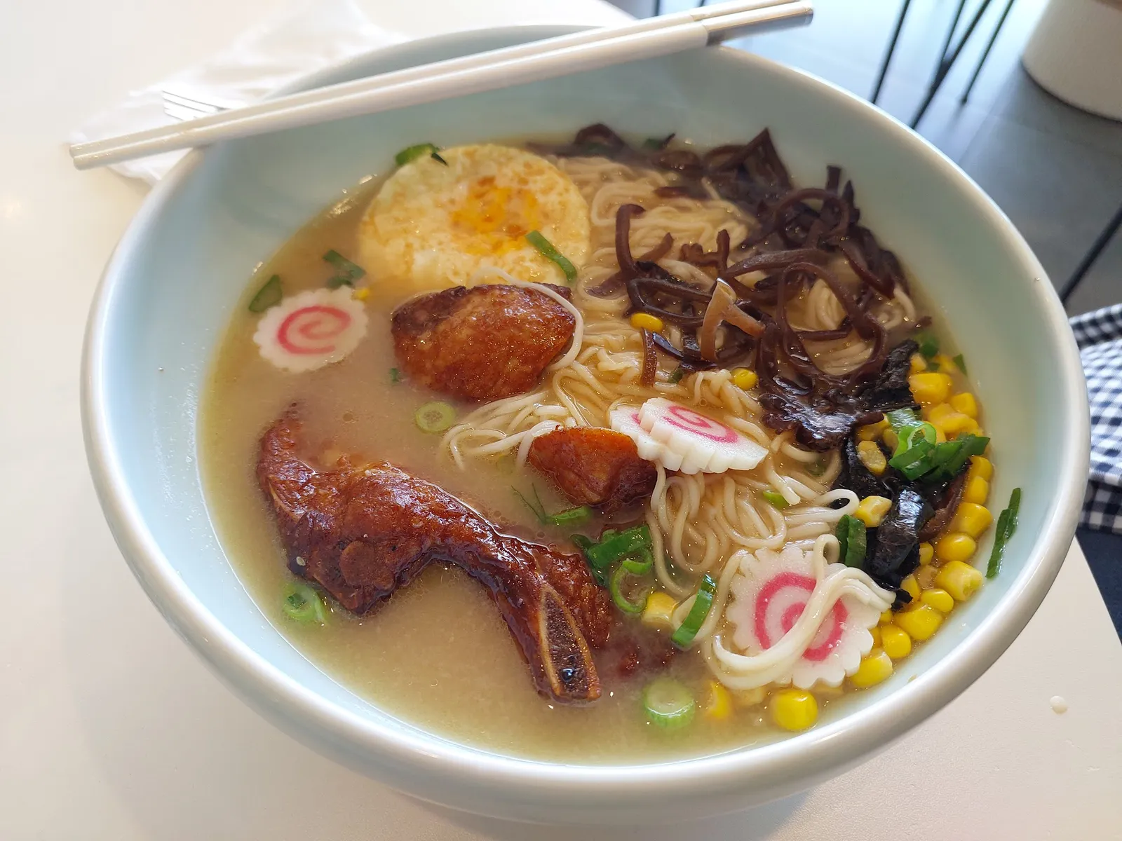 Overhead view of pale-blue bowl of pork rib ramen with egg, rib pieces, corn and spring onion