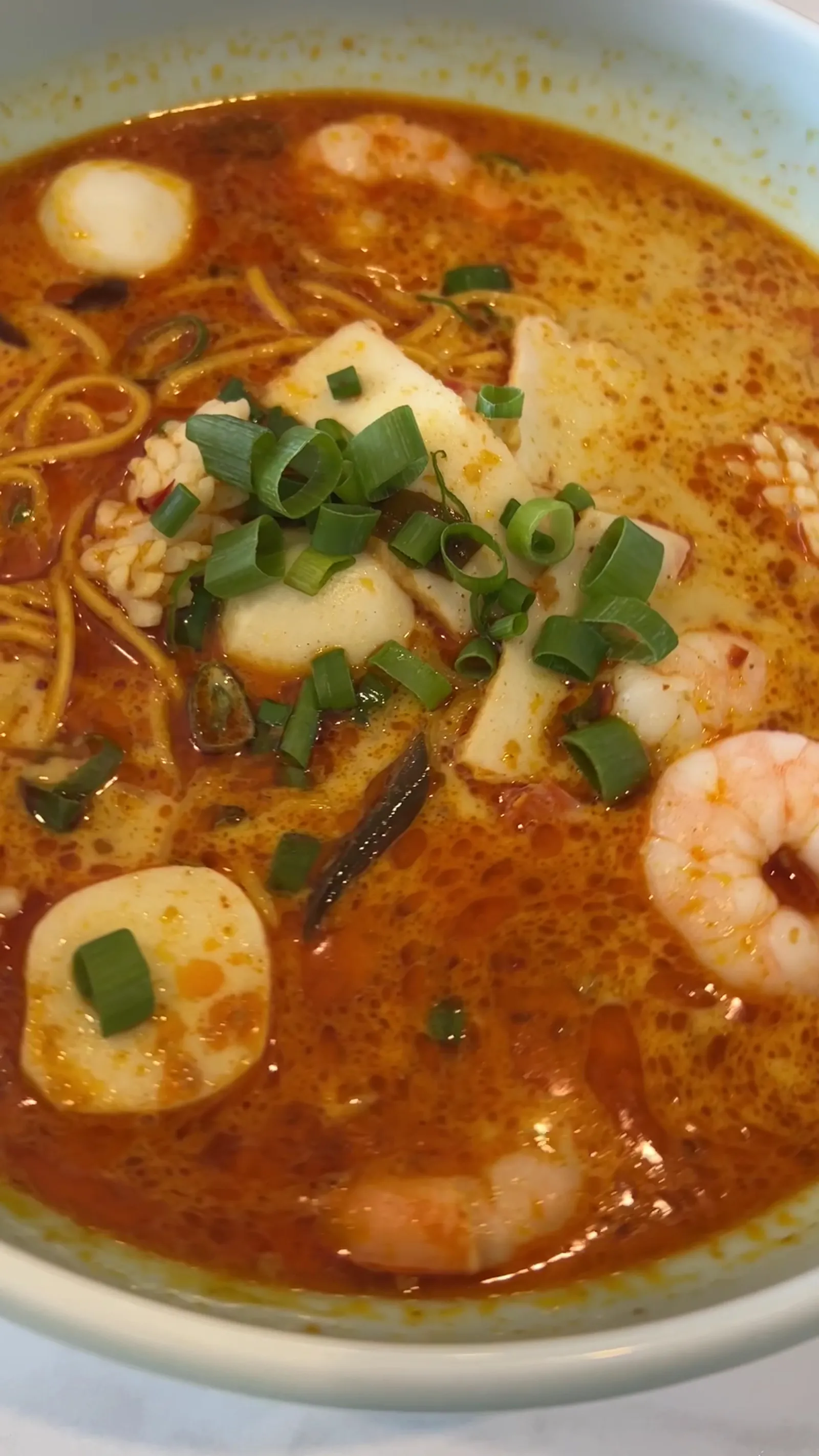 Close-up of spicy tomyum ramen with prawns, fish tofu, narutomaki and spring onion in red-orange broth