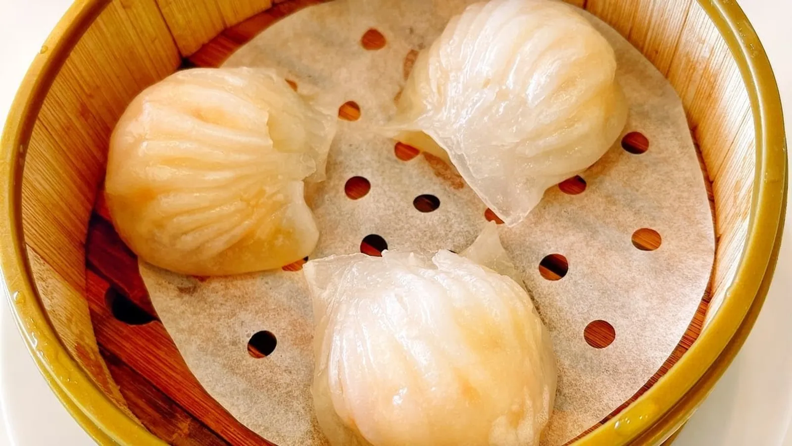 Three glistening steamed prawn dumplings in bamboo steamer basket on white parchment paper